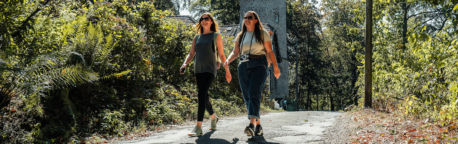 Zwei Frauen mit Sonnenbrillen wandern einen Urban Trail. Im Hintergrund steht ein historischer Steinturm.