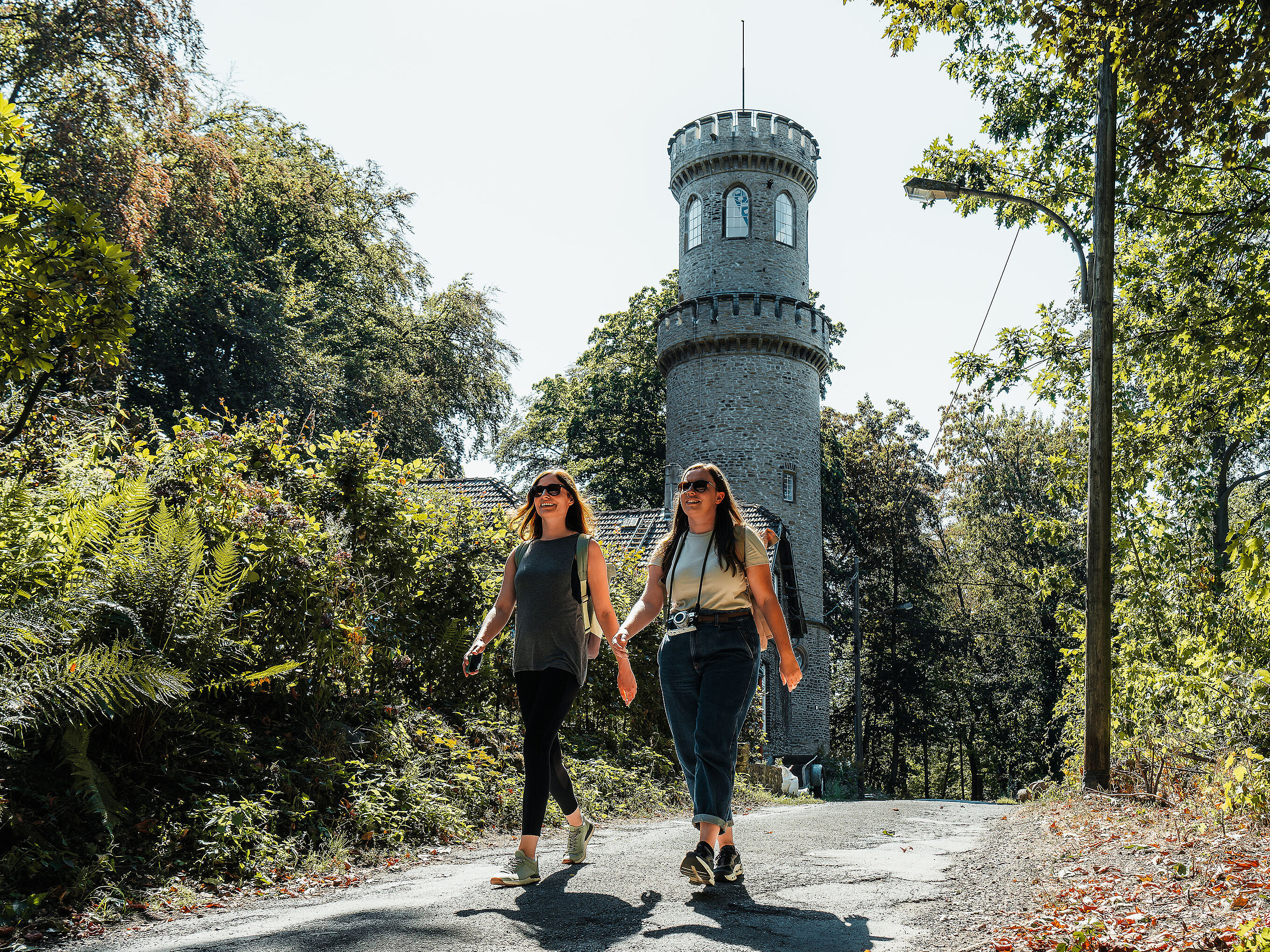 Zwei Frauen mit Sonnenbrillen wandern einen Urban Trail. Im Hintergrund steht ein historischer Steinturm.
