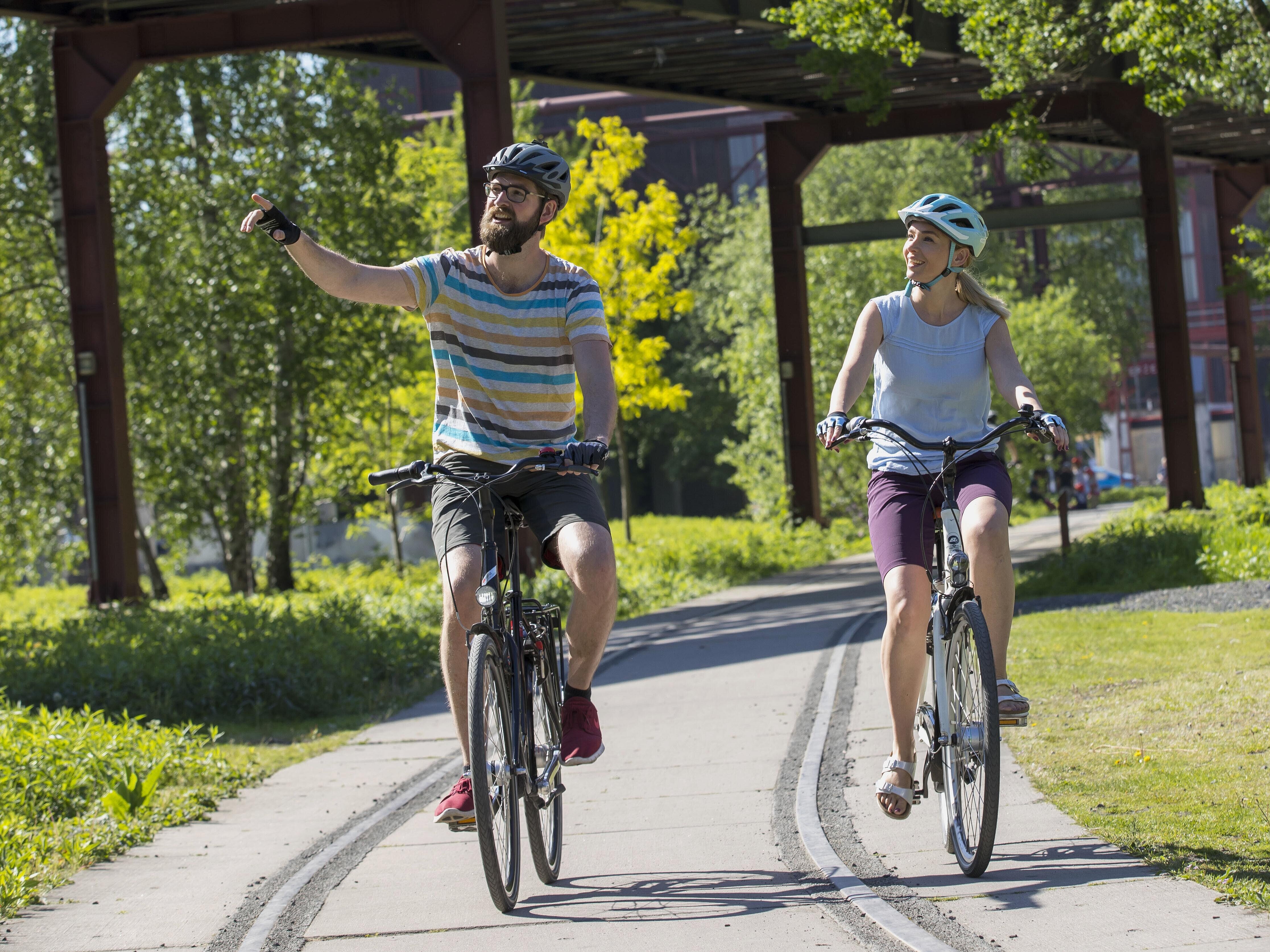 Ein erwachsenes Pärchen fährt mit dem Fahrrad auf der Radrevier Route, im Hintergrund ist das Weltkulturerbe Zeche Zollverein zu sehen.